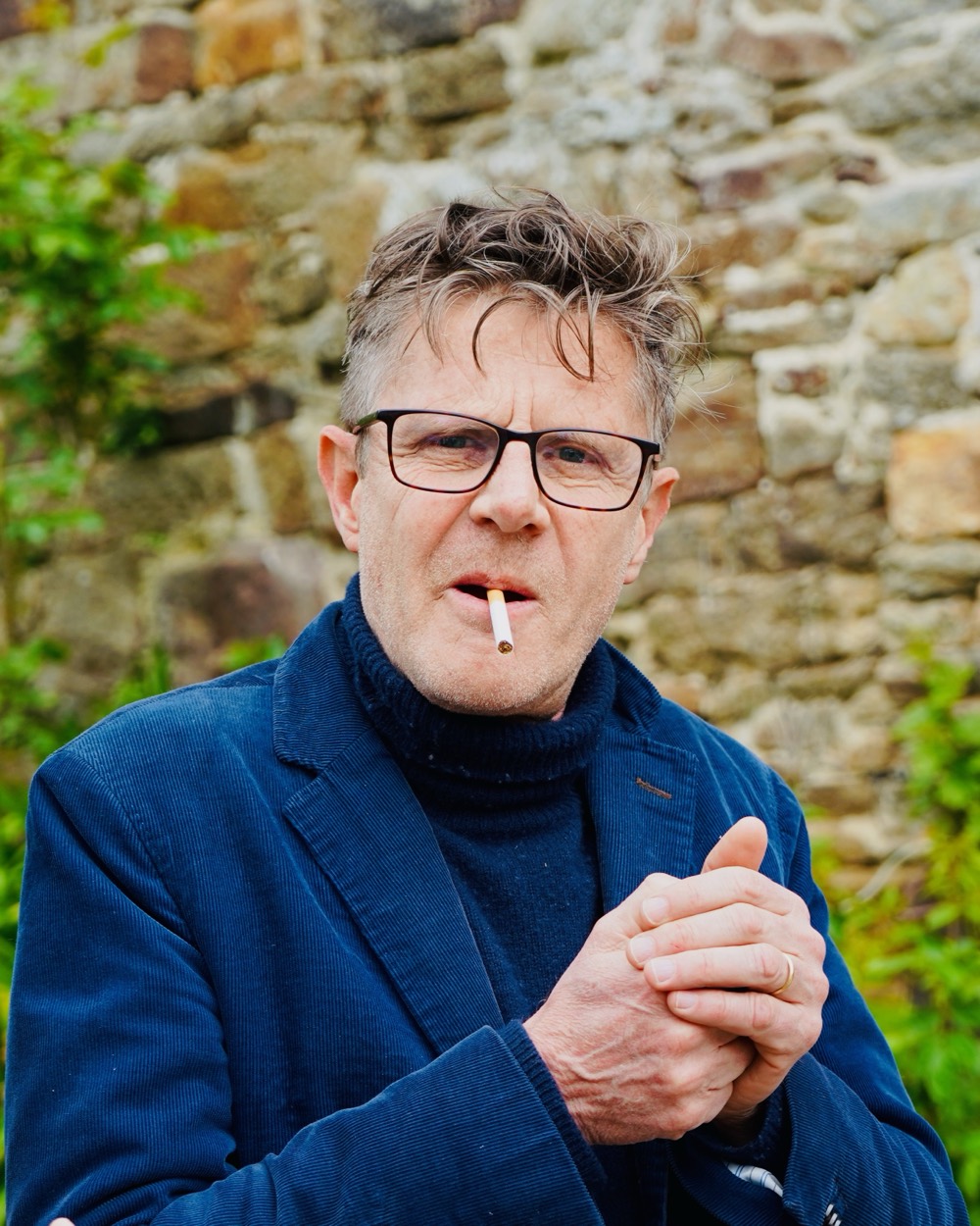 Portrait d'un homme à lunettes, veste bleue, cigarette aux lèvres, mur de pierre